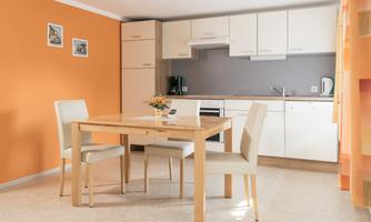 A modern kitchen with a dining table and four chairs. The walls are painted orange and there are light-colored furniture.