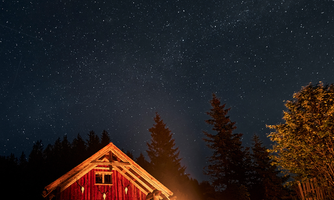 A cozy campfire in front of a wooden barn under a clear starry sky. People sit around the fire and enjoy the night.
