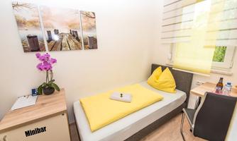 A simple room with a single bed and yellow cushions. On the table, there is a small minibar and a flower, and there is a window with yellow curtains.