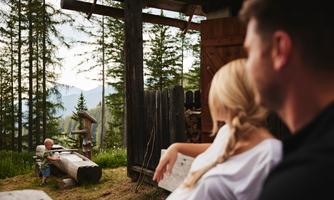 A family is sitting relaxed in front of a cabin in nature. In the background, a small child is playing with a tree trunk.