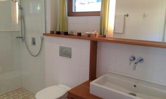 A modern bathroom with a shower and a wooden sink installation. There are bright windows with curtains and minimalist design.