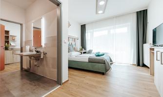A modern room with a double bed and plenty of natural light. In the background, a television and an open bathroom are visible.