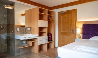 A modern bedroom with a double bed and warm wooden elements. The room features a sink and an open wardrobe.
