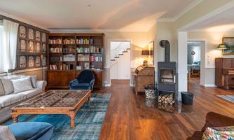 A cozy living room with a bookshelf, a comfortable sofa, and a wood stove. The room has beautiful wooden floors and is bright with natural light.