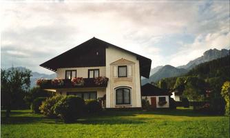 A pretty house with a large garden and colorful flowers. In the background, there are mountains and a cloudy sky.