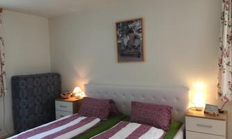 A cozy bedroom with two single beds and a bright, friendly atmosphere. Pictures hang on the walls, and the windows are adorned with floral curtains.