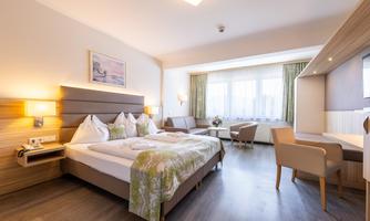 A modern hotel room with a double bed and two pillows. There is a small living area with a table and chairs as well as large windows that let in daylight.