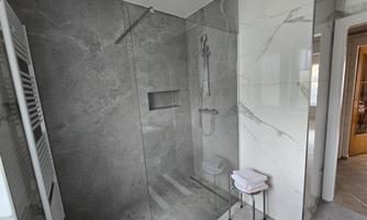 A modern shower with gray tiles and a clear glass pane. Towels are on a small table.