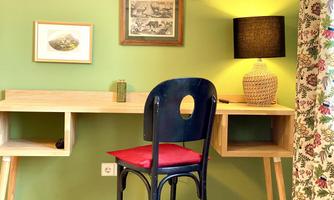 A cozy workspace with a wooden desk and a blue chair with a red cushion. In the background, there is a picture, a lamp, and a decorative curtain.