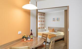 A cozy living space with a dining table and chairs. In the background, there is a bedroom with a double bed and orange accents.