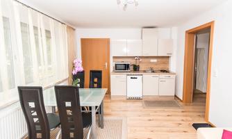 A modern kitchen with a dining area and large windows. The decor is bright and inviting, featuring a table and black chairs.