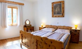 A bright bedroom with a wooden double bed and an elegant bedside table. The room has large windows with curtains and a cozy atmosphere.