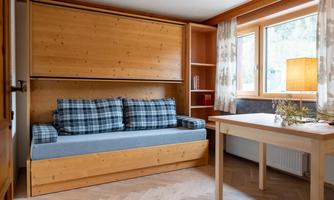 A cozy room with a foldable sleeper sofa and a wooden shelf. A table and a window with curtains create a bright atmosphere.