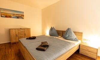 A cozy bedroom with a large bed and modern wooden furniture. The walls are painted light, and a picture hangs above the bed.