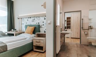 A modern hotel room with a comfortable bed and a cozy armchair. In the background, a bathroom with white tiles and wooden shelves can be seen.