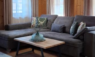 A cozy living room with a gray sofa and a wooden table. Windows with curtains create a bright, inviting atmosphere.