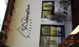 A modern building with the inscription "Alpenflair" and stylized mountains. In front of the house, there is a small garden with plants and furniture.