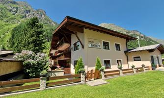 A cozy house in the mountains with a green garden. Surrounded by trees and a beautiful mountain backdrop.
