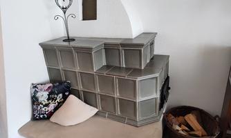 A modern tile stove in a corner of a room. Next to it is a cozy bench with a cushion and a basket of wood.