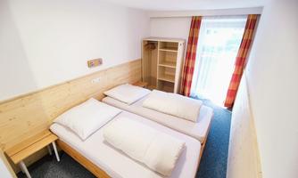 A bright room with two single beds, wooden shelves, and large windows. The warm color scheme creates an inviting atmosphere.