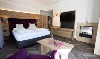 A modern hotel room with a double bed and a cozy seating area. The room is bright and stylishly furnished, featuring a television and a fireplace.