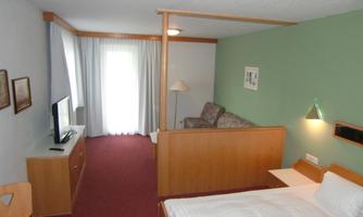 A spacious hotel room with a double bed and a separate living area. It features a television, a desk, and a large window with curtains.