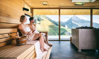 A couple relaxes in a modern sauna with a view of the mountains. Large windows provide a stunning view of the green landscape.