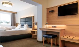 A modern hotel room with a double bed, a table, and a television. The furnishings are made of light wood and create a pleasant atmosphere.