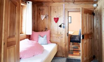 A cozy room with wooden walls and a bed with checkered bedding. In the background, a dining table is visible and the door is open.
