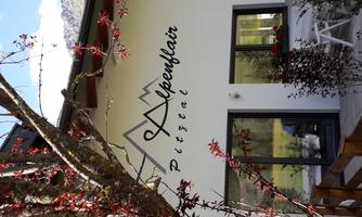 A modern building with the inscription "Alpenflair Pitztal". The surroundings are surrounded by mountains and trees.