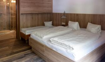 A simple bedroom with two single beds and wooden paneling. In the background, a modern bathroom is visible.