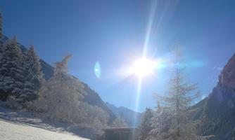 A snowy landscape with high mountains in the background. The sun shines brightly in the clear blue sky.