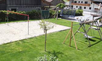 A well-maintained garden with a sand volleyball court and a playground. In the background, residential houses and a green landscape can be seen.
