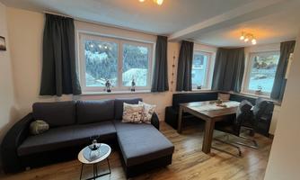 A cozy living room with a modern couch and a dining area. Large windows provide a view of the snowy landscape.