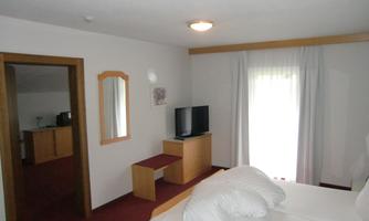 A simple and bright bedroom with two beds, a television, and a table. Daylight shines in through the windows.