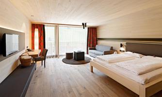 A modern hotel room with wooden floors and large windows. It features a double bed, a seating area, and a desk.
