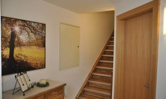 A bright hallway with a wooden staircase and a painting of a sunny forest. A small table with decorations stands against the wall.