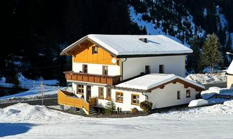 A cozy house in the snow with a bright wooden roof.  
Surrounded by an impressive mountain landscape and a radiant blue sky.