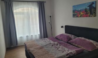 A cozy bedroom with a double bed and purple bedding. Large windows let in plenty of natural light.