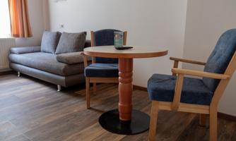 A cozy living space with a gray sofa, a round table, and two chairs. The windows are adorned with orange curtains.