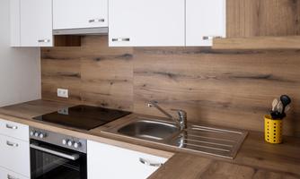 A modern kitchen with white cabinets and wood look. The work area includes a cooktop, a sink, and kitchen utensils in a yellow container.