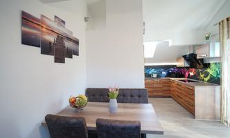 A modern kitchen with wooden furniture and a cozy dining area. Pictures of a landscape hang on the wall, and the room is bright and friendly.