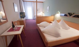A cozy hotel room with a double bed and large pillows. A table with a bottle of wine and a glass is next to the bed.