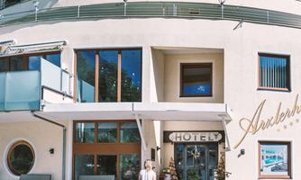 A person with a suitcase stands in front of the hotel "Auhof".  
The building has several floors and large windows, surrounded by a clear blue sky.