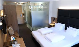 A modern hotel room with a double bed and two pillows. In the background, a glass bathroom is visible.