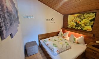 A cozy bedroom with a double bed and colorful cushions. On the wall, there are pictures of flowers and a wooden ceiling visible.