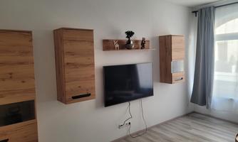 A modern living room with a flat-screen TV on the wall. The furniture is made of light wood and there is a large window area with a curtain.