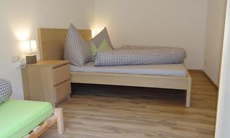 A simple bedroom with two beds and a nightstand. The floor is made of wood and the walls are painted light.