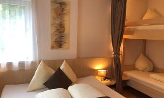 A cozy bedroom with a double bed and a bunk bed in the corner. The windows let in plenty of daylight and there are decorative cushions and a lamp.