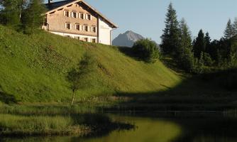 A cozy house on a gentle hill with a clear lake in the foreground. In the background, trees and a mountain can be seen.